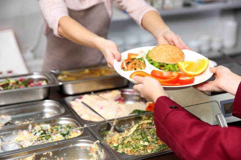 A cafeteria worker hands a plate of food to a student across a school lunch counter.
