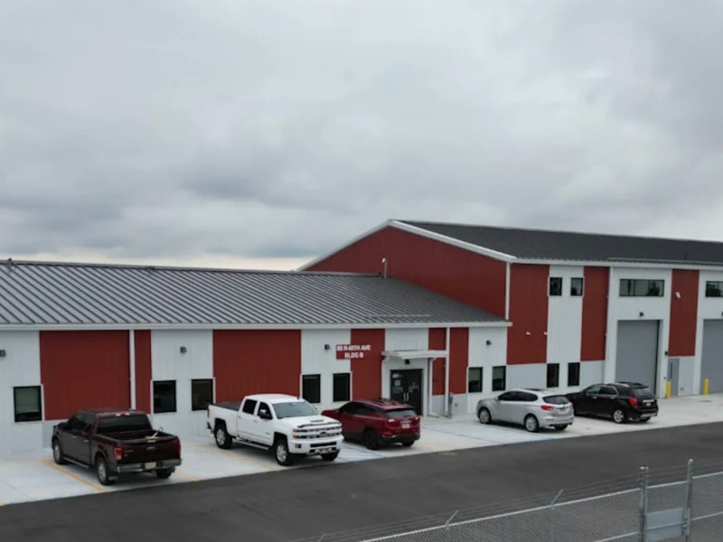 A wide view of the red and white exterior of 27J Schools’ new North Transportation Terminal in Brighton, Colorado. Several vehicles are parked in front of the building, which features large bay doors and office entrances under an overcast sky.