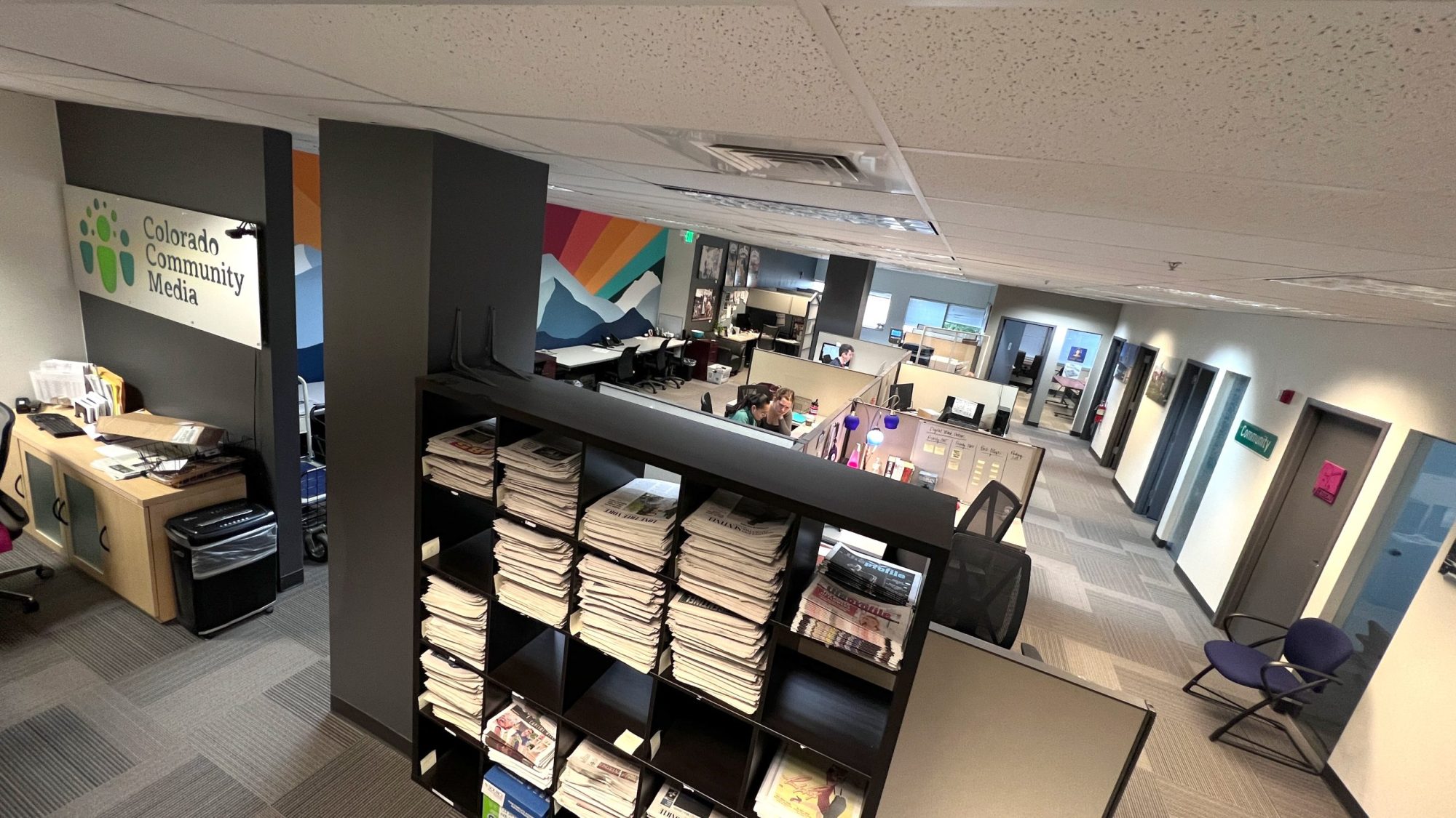 Aerial view of a newspaper office. Stacks of newspapers in a black credenza. Reporters working t their desk in the background.