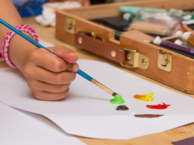 Close-up of a child's hand painting with green acrylic on paper, with a wooden art supply box in the background.