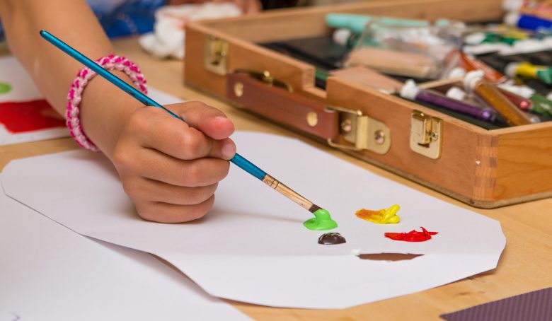 Close-up of a child's hand painting with green acrylic on paper, with a wooden art supply box in the background.