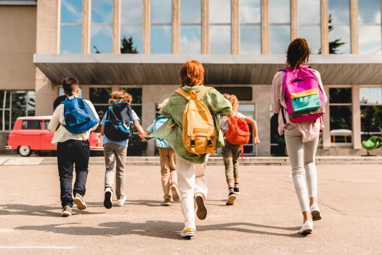Group of elementary school children wearing backpacks and walking toward a school building, symbolizing the return to school and the impact of education funding changes.