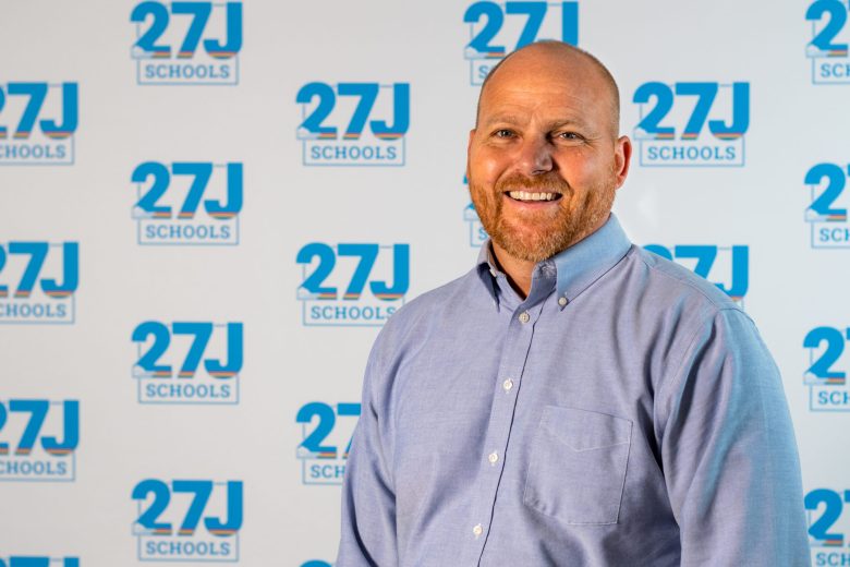 Will Pierce, smiling in a light blue button-down shirt, stands in front of a 27J Schools logo backdrop.
