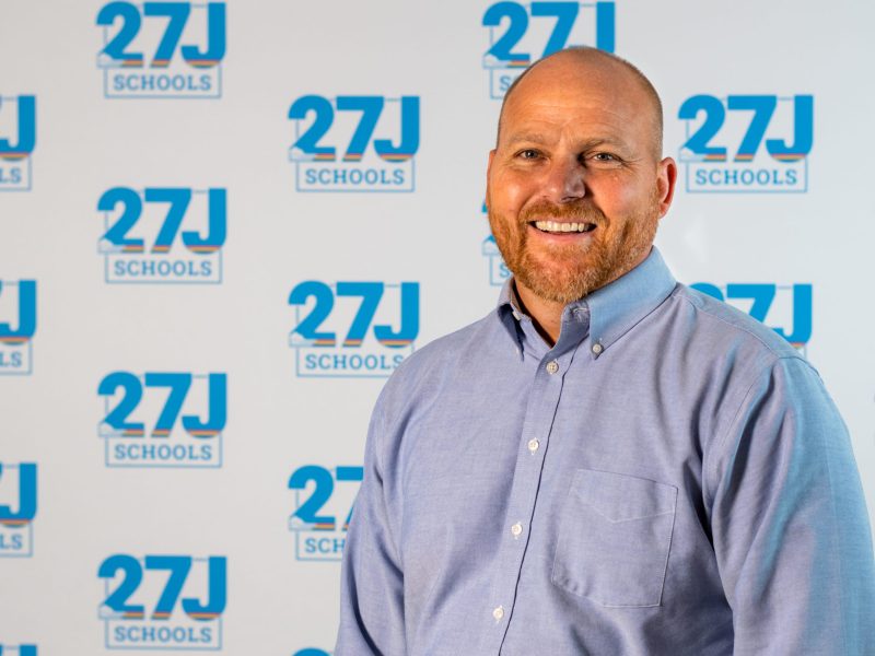 Will Pierce, smiling in a light blue button-down shirt, stands in front of a 27J Schools logo backdrop.